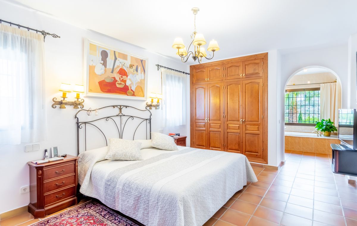 Dormitorio con cabecero de forja, armarios empotrados de madera y suelo de terracota. Un arco conecta con el baño en suite que incluye bañera y vistas exteriores.