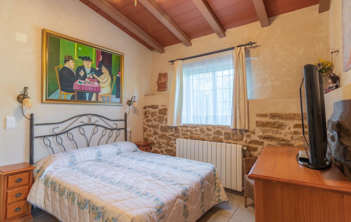 Dormitorio rústico con cama de forja, paredes de piedra vista y techos de vigas de madera. Incluye un gran cuadro figurativo y mobiliario de madera clásica.