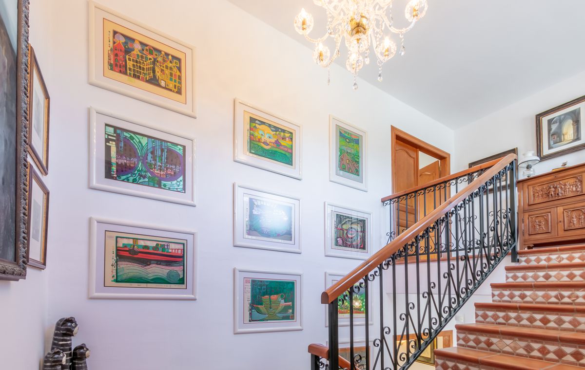 Escalera con peldaños de terracota y contrahuellas decoradas, barandilla de hierro forjado y una pared de galería con obras de arte bajo una lámpara de araña.