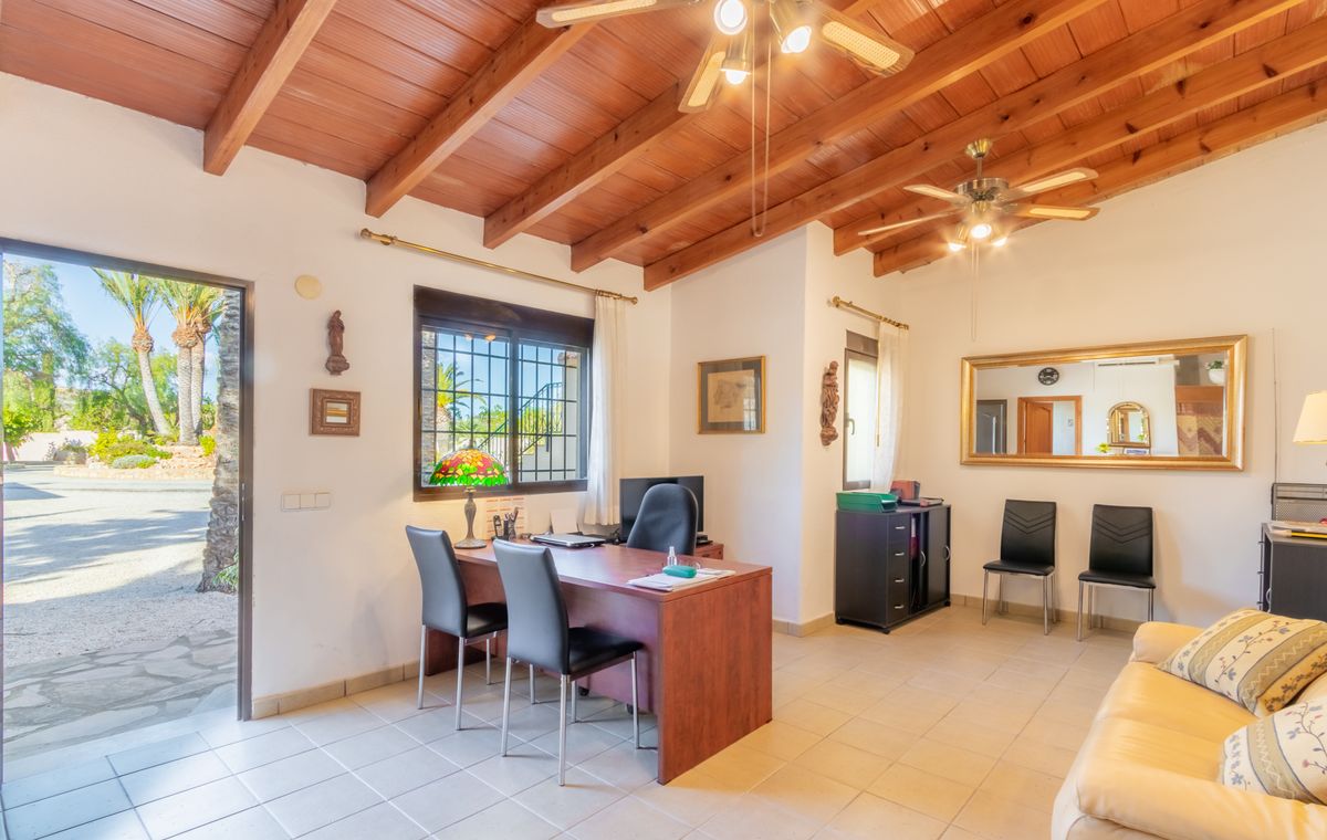 Oficina con techo de vigas de madera, ventiladores y escritorio. La puerta abierta conecta con el exterior, mostrando palmeras y luz natural sobre el suelo cerámico.