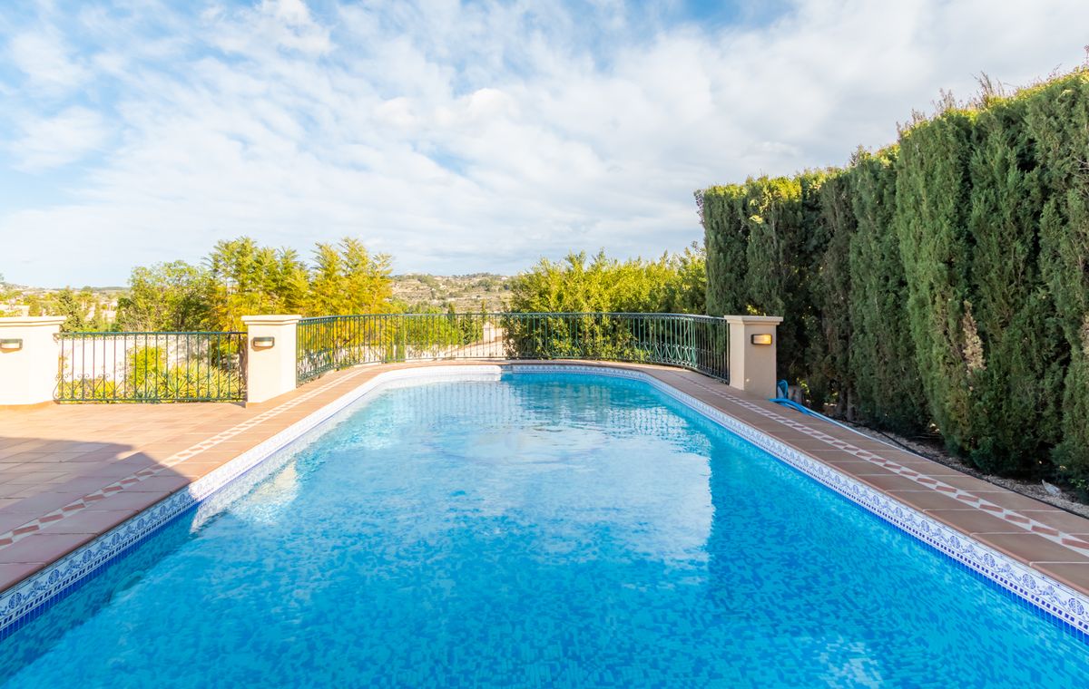 Piscina exterior con revestimiento de mosaico azul, borde de terracota decorado y una valla de forja negra junto a un seto alto de cipreses.