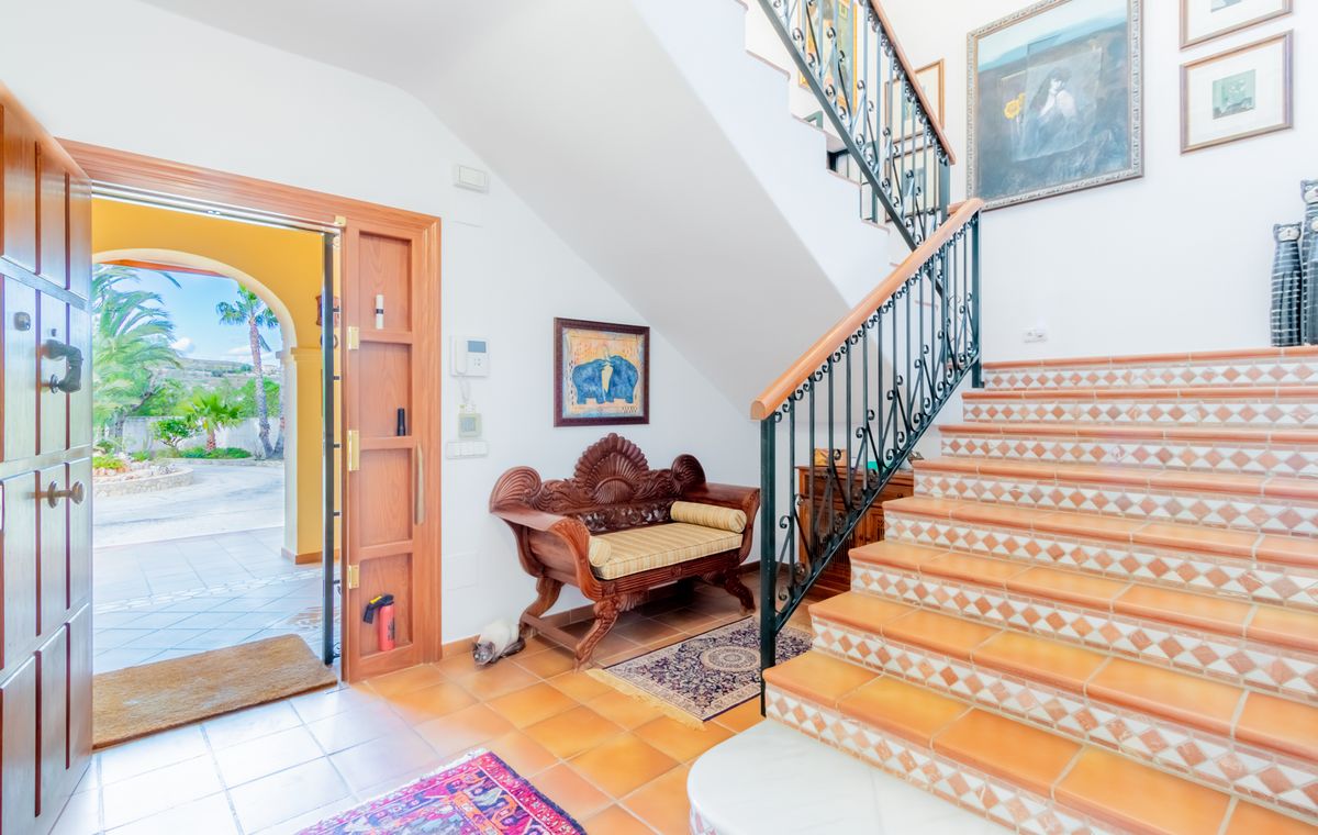 Recibidor con escalera de baldosas de terracota y contrahuellas decorativas, barandilla de hierro forjado y un banco de madera tallada. La puerta abierta da paso al patio exterior.