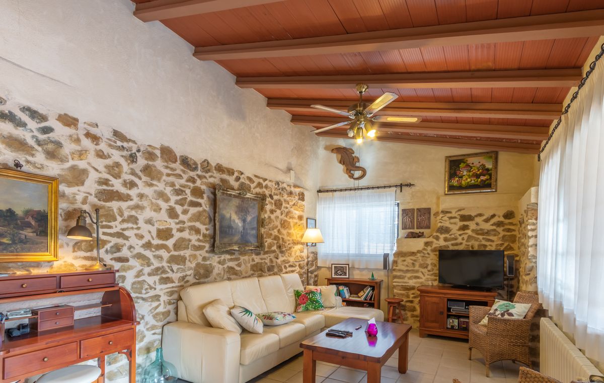 Sala de estar rústica con paredes de piedra natural vista, techos de vigas de madera rojiza y un sofá de cuero crema junto a un escritorio clásico.