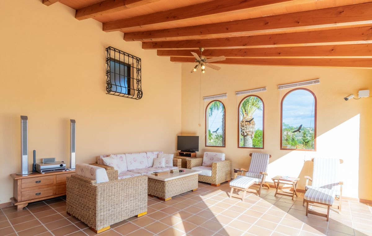 Salón acristalado con vigas de madera vistas, suelo de terracota y muebles de mimbre. Tres ventanas de arco ofrecen luz natural y vistas a las palmeras.