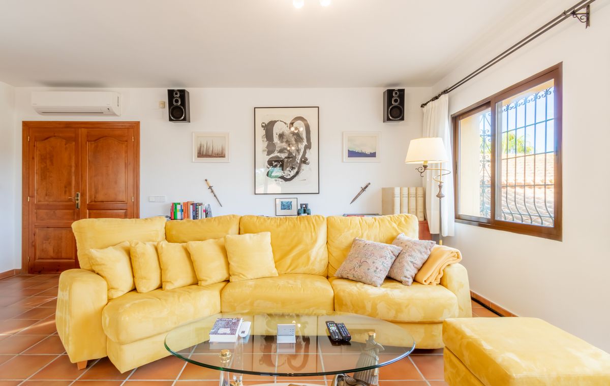 Salón con un amplio sofá rinconera amarillo, mesa de centro de cristal y suelo de terracota. Las paredes blancas incluyen obras de arte y altavoces integrados.