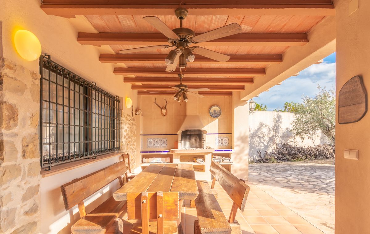 Terraza cubierta con techo de vigas de madera y ventiladores, zona de comedor rústica y barbacoa de obra decorada con azulejos tradicionales y piedra.