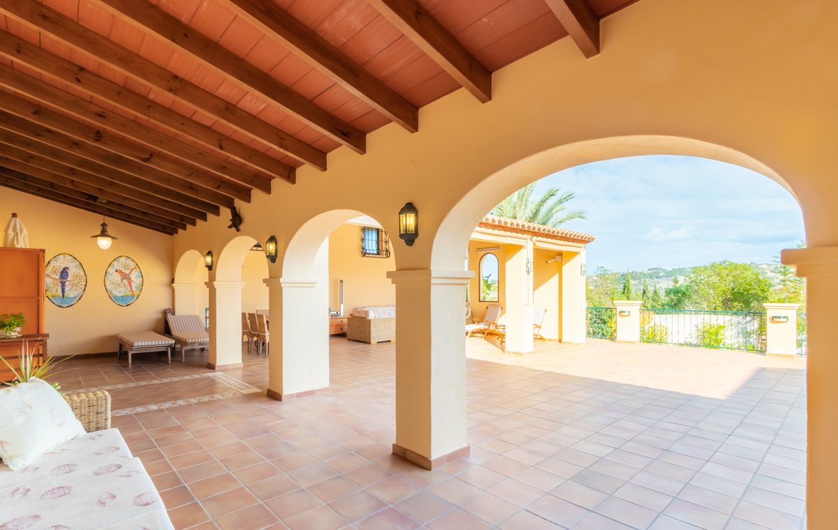 Terraza cubierta con vigas de madera oscura, suelo de baldosas de barro con cenefas decorativas y arcos que conectan con un amplio patio exterior.