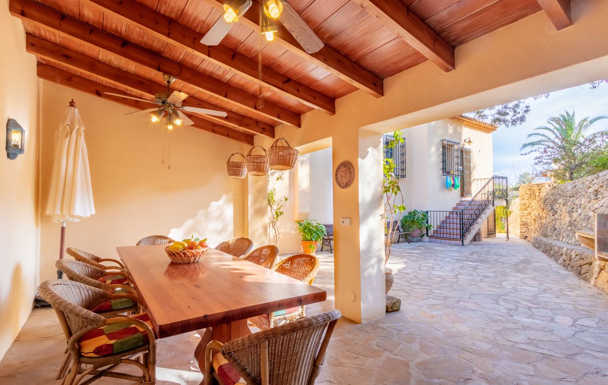 Terraza cubierta con vigas de madera, ventiladores de techo y zona de comedor con sillas de mimbre, abierta a un patio empedrado tradicional.