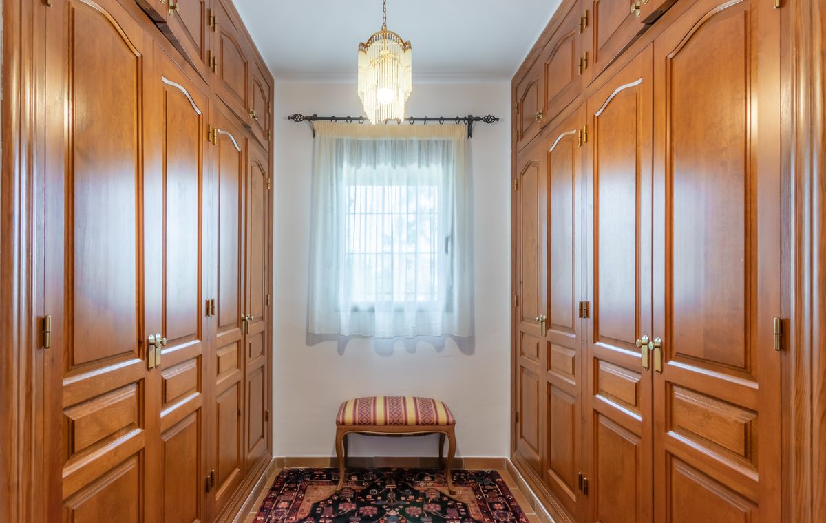 Vestidor con amplios armarios empotrados de madera, lámpara de araña clásica y ventana central con luz natural.