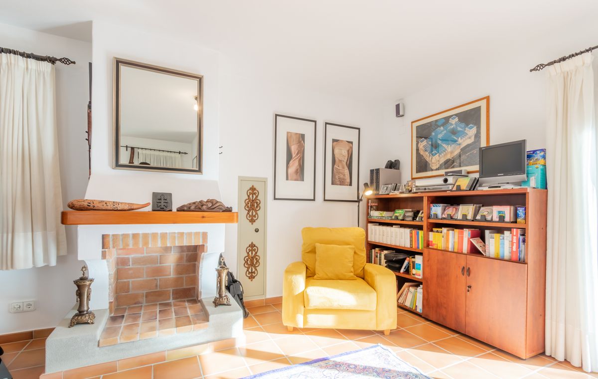 Zona de estar con chimenea de ladrillo visto, sillón amarillo y estantería de madera sobre suelos de baldosa de terracota.