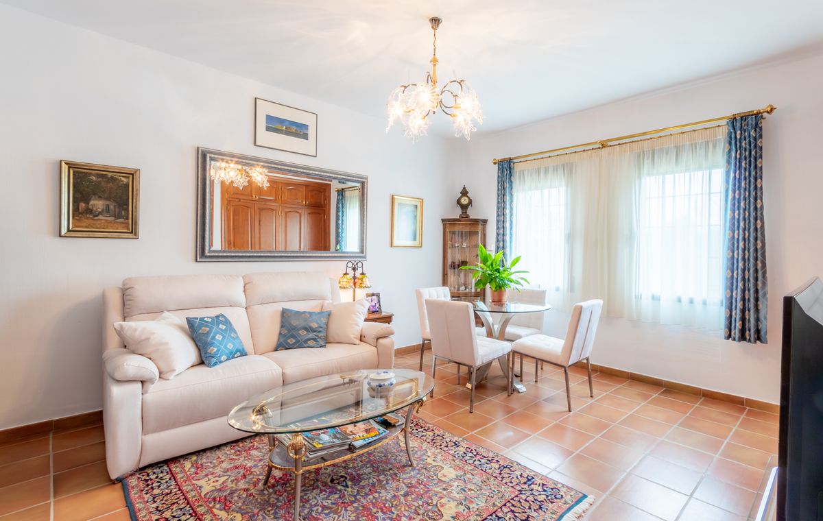 Zona de estar luminosa con suelos de baldosa de terracota, sofá blanco con cojines azules y conjunto de comedor de cristal bajo una lámpara ornamentada.