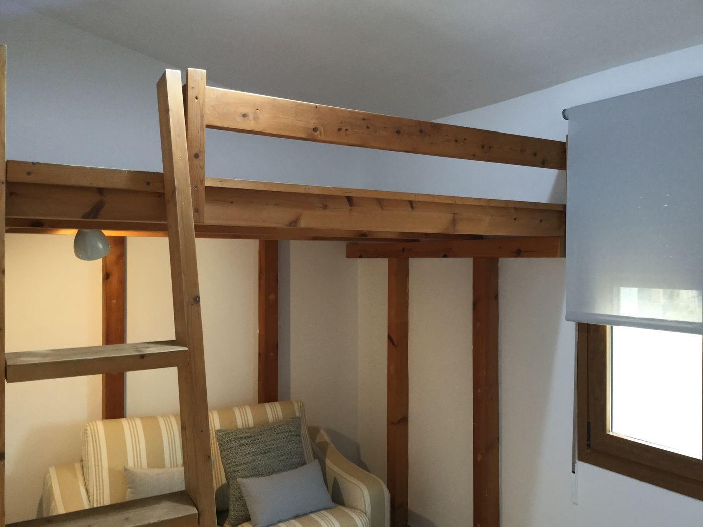 Altillo con estructura de cama elevada en madera maciza y escalera. Incluye una zona de estar inferior con un sofá de rayas y una ventana con estor enrollable gris.