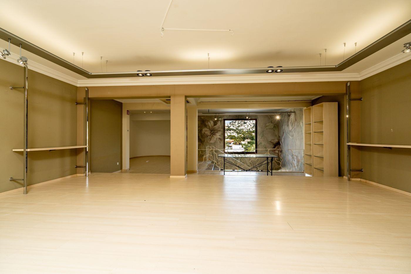 Amplio local comercial con suelos de madera clara, iluminación indirecta en el techo y soportes cromados. Al fondo destaca un mural floral y una ventana con vistas exteriores.