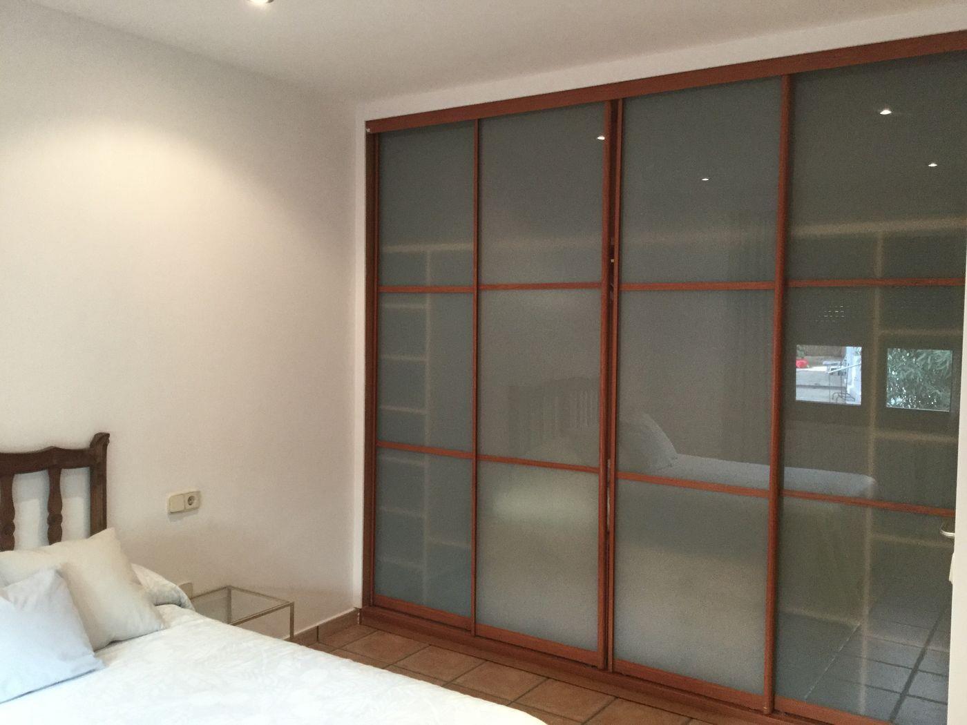 Armario empotrado de cuatro puertas correderas de cristal translúcido y marcos de madera. El dormitorio incluye suelos de baldosa cerámica y focos empotrados.
