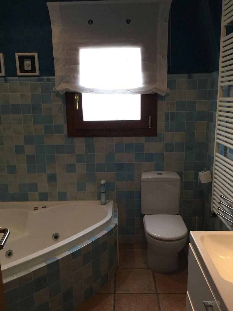 Baño con bañera de hidromasaje rinconera, azulejos de mosaico en tonos azules y radiador toallero blanco. Ventana de madera oscura con luz natural.