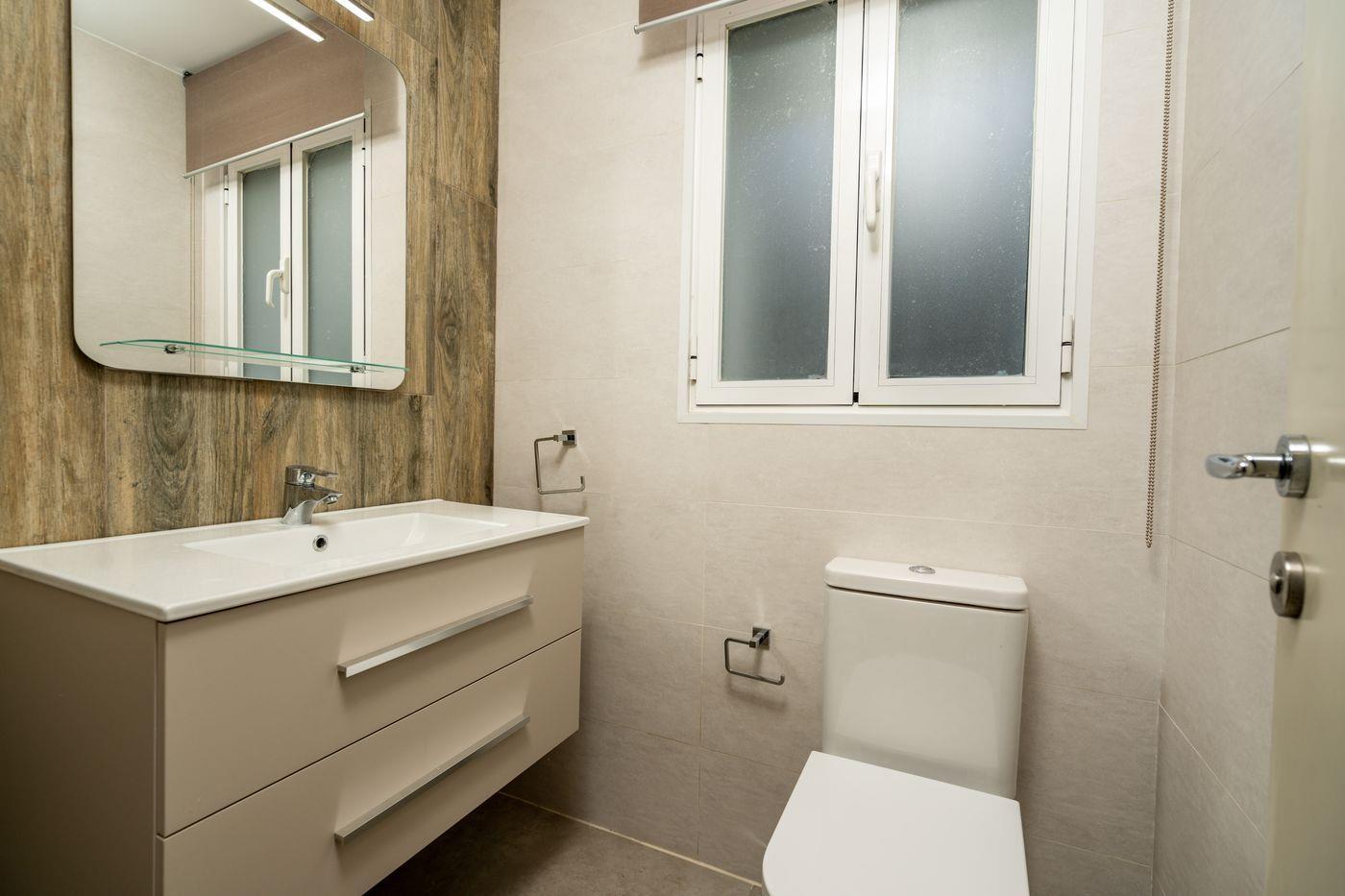 Baño moderno con mueble blanco de cajones, pared de acento con efecto madera, espejo amplio y ventana de cristal translúcido.