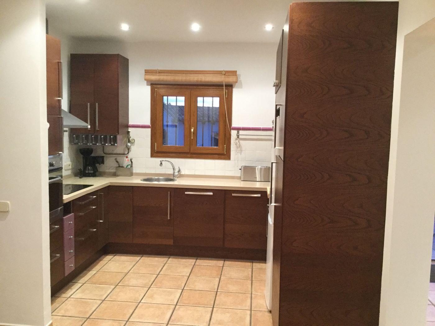 Cocina funcional con armarios de madera oscura, encimera clara y suelos cerámicos. Incluye una ventana de madera con persiana de bambú e iluminación empotrada.