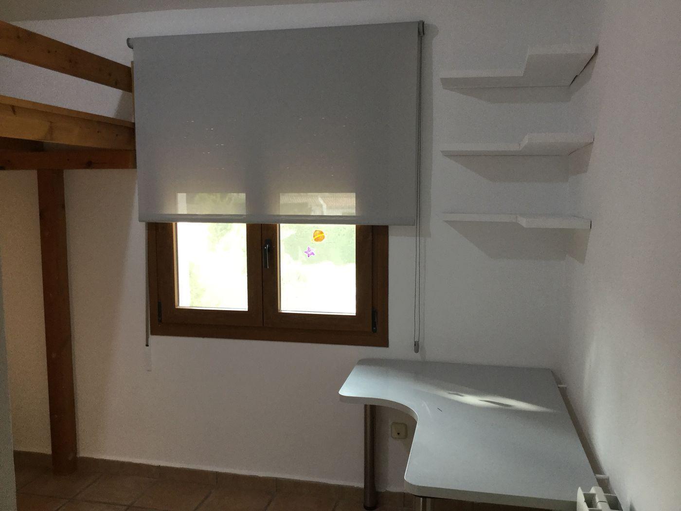 Dormitorio con cama alta de madera, escritorio de rincón y estanterías flotantes blancas. La ventana de madera incluye un estor enrollable gris para regular la luz.