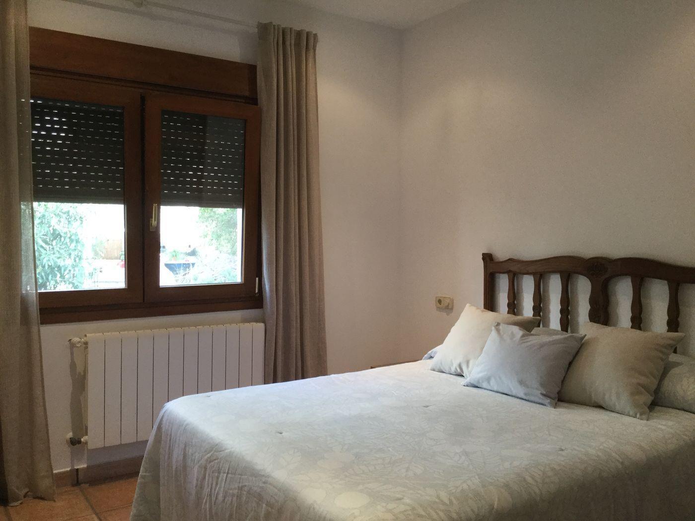 Dormitorio con cama doble, cabecero de madera tallada, ventana de madera con persianas integradas y radiador de pared bajo el ventanal.