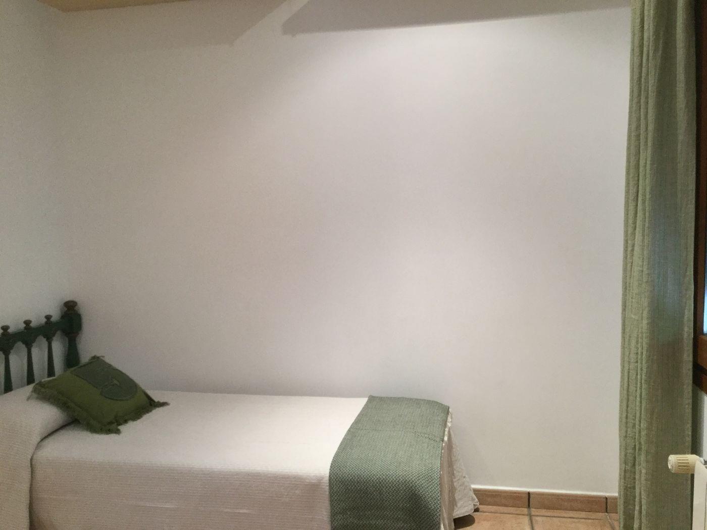 Dormitorio individual con cabecero de madera oscura, ropa de cama blanca con detalles en verde, suelo de baldosas de terracota y cortina a juego.