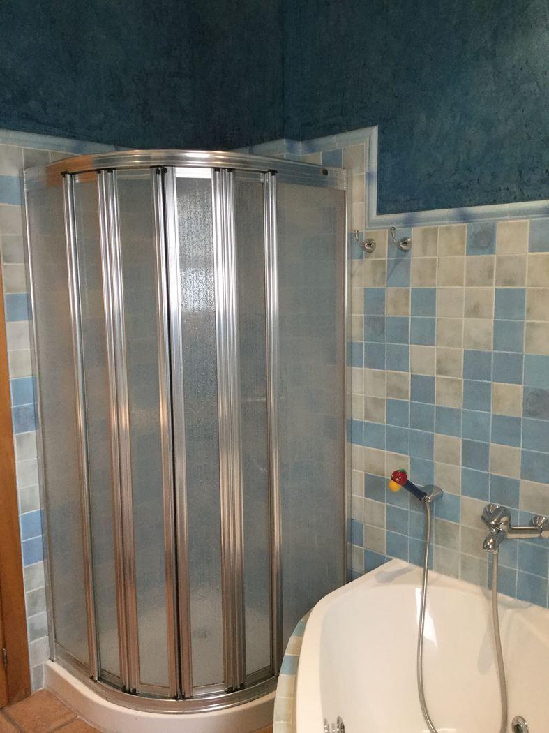 Ducha de esquina con mampara curva de aluminio y cristal translúcido, junto a una pared de mosaicos azules y beige y una bañera blanca.