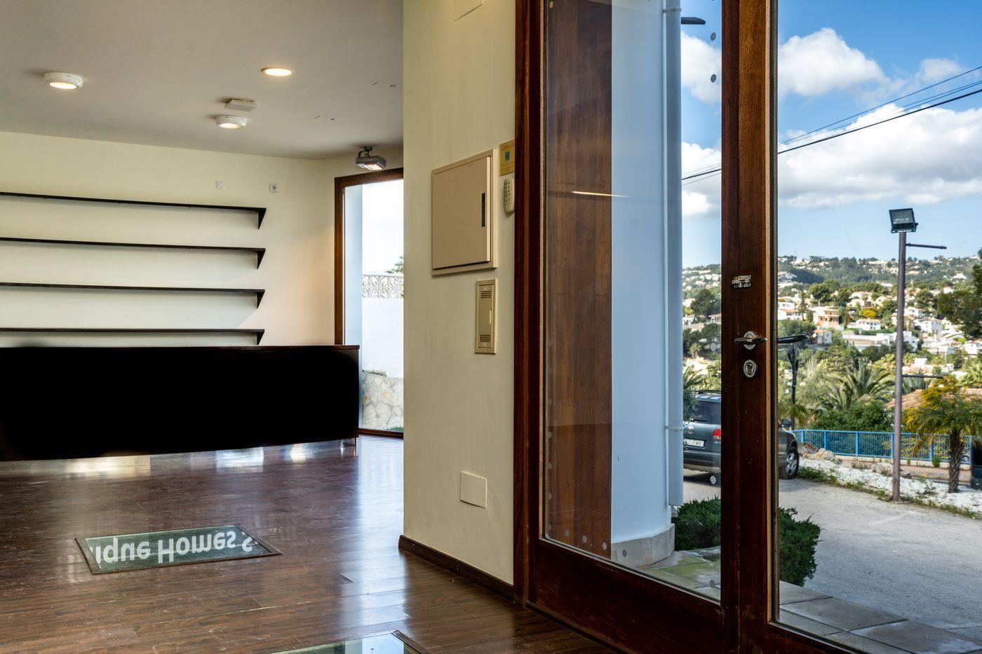 Entrada con suelo de madera oscura, mostrador de recepción negro y puerta de cristal. La vista muestra una zona residencial montañosa con casas blancas y cielo despejado.