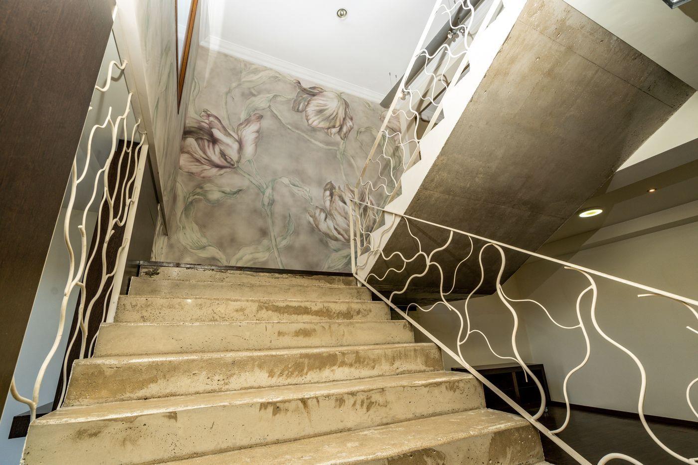 Escalera de hormigón visto con barandilla de hierro blanco ondulado, destacando una pared con papel pintado de tulipanes y focos empotrados.