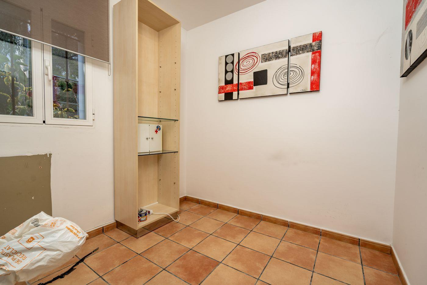 Habitación interior con suelo de baldosas de terracota, una estantería alta de madera clara con baldas de cristal y cuadros abstractos en la pared.