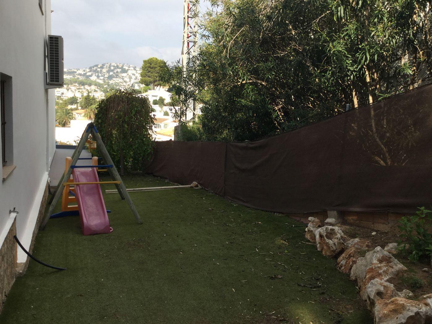 Jardín lateral con césped artificial y zona de juegos infantiles. Incluye un tobogán y columpio, protegidos por una malla de ocultación con vistas al paisaje urbano.