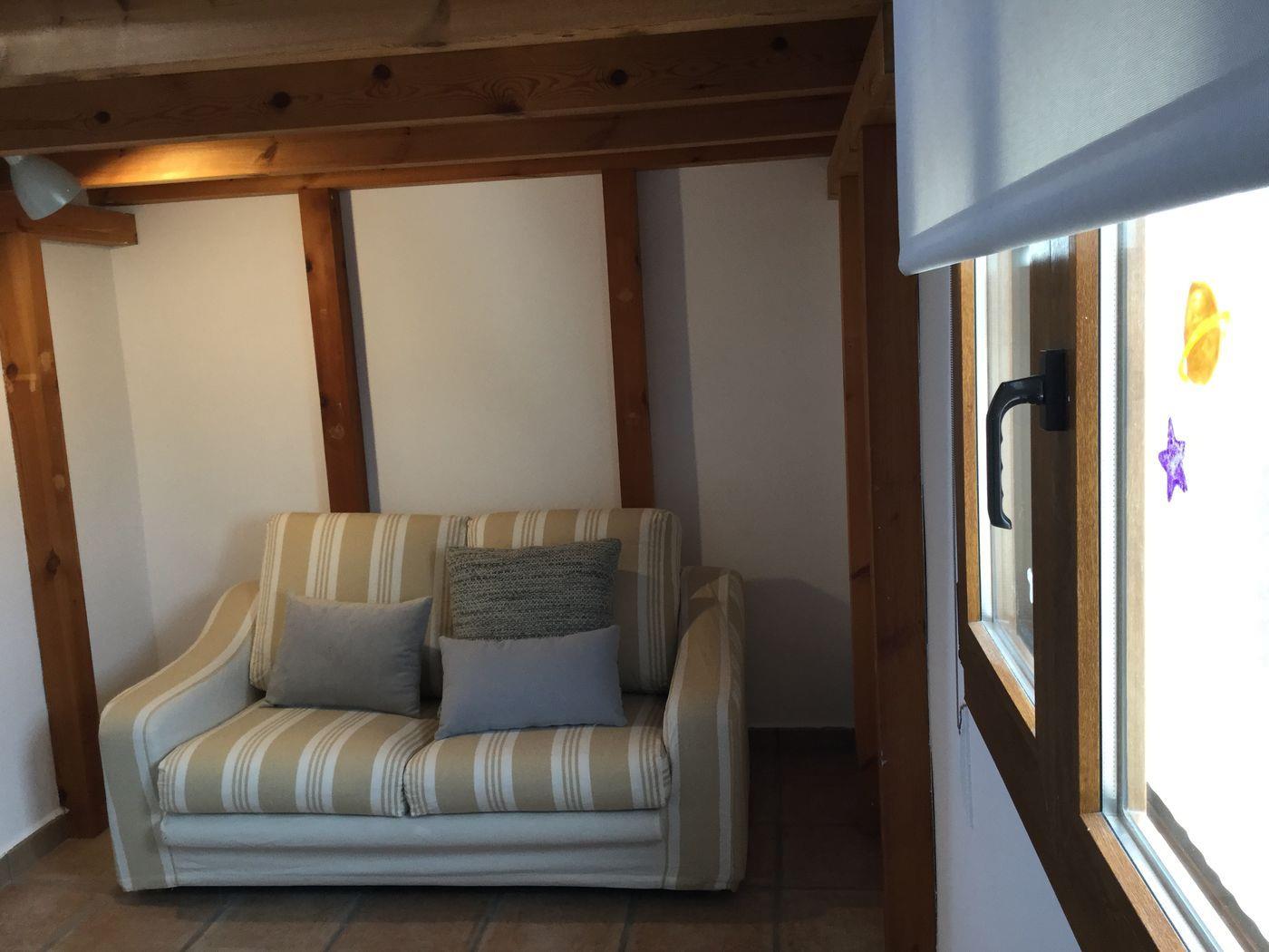 Rincón de descanso con sofá de rayas y vigas de madera vista. Una ventana con marco de madera aporta luz natural a esta estancia de estilo rústico.