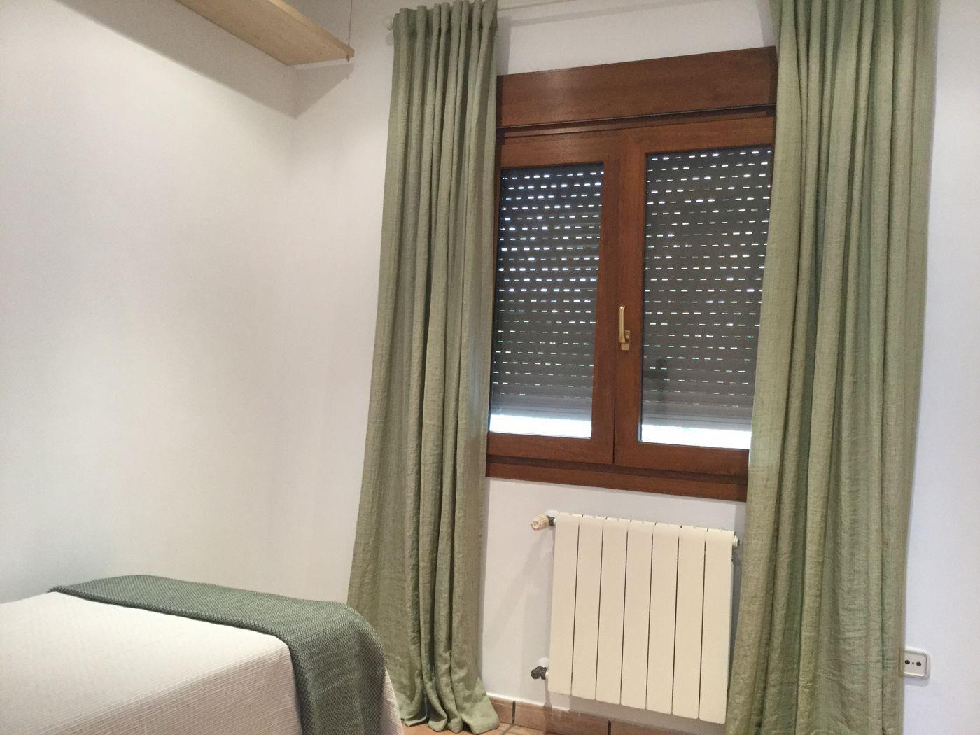 Rincón de dormitorio con ventana de madera y persianas integradas, cortinas largas en verde salvia y radiador blanco. Incluye cama con manta a juego y paredes blancas.