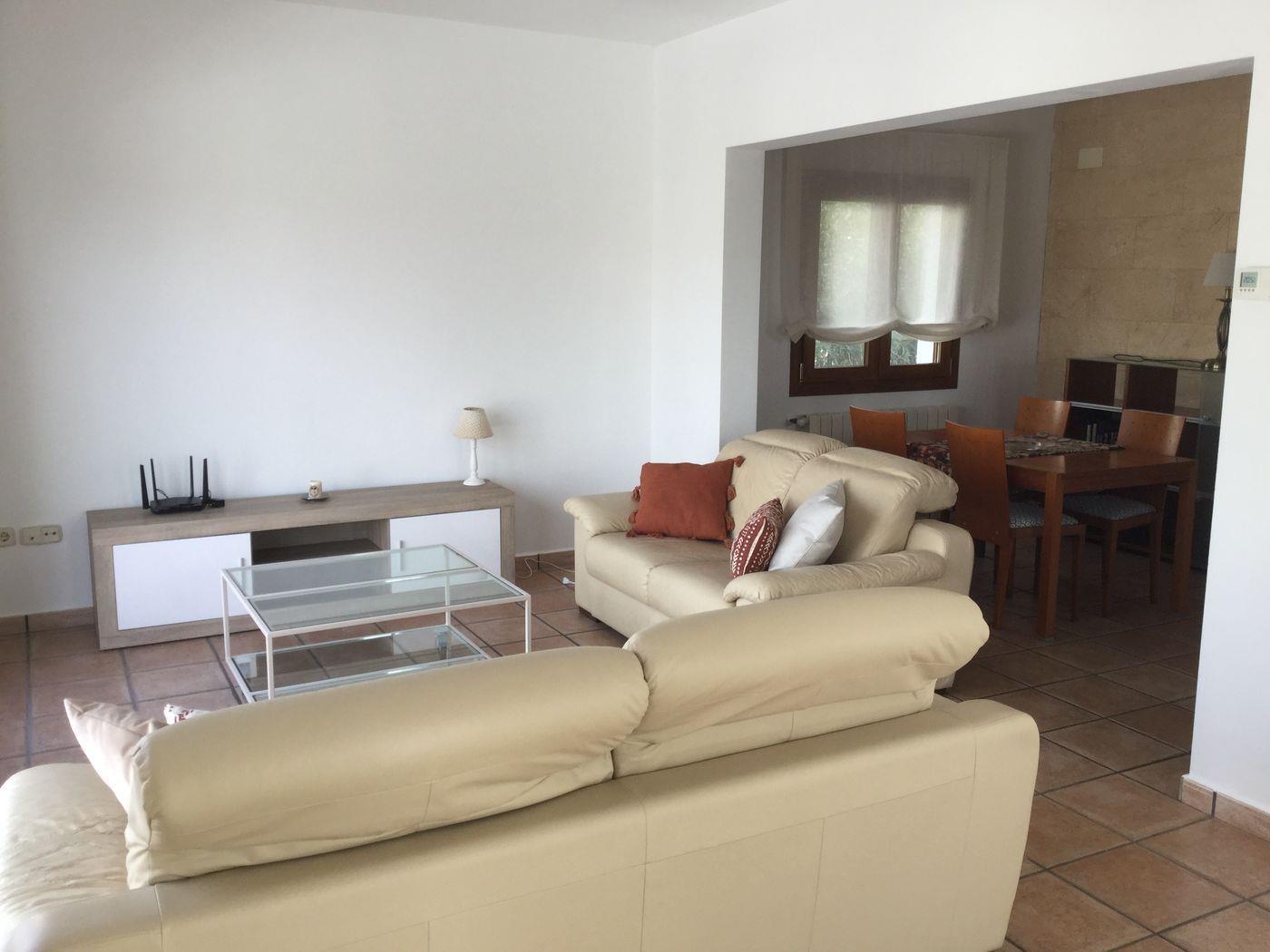 Salón comedor con sofás de cuero crema, mesa de centro de cristal y suelos de gres rústico. Al fondo se aprecia el comedor con una pared de piedra natural.