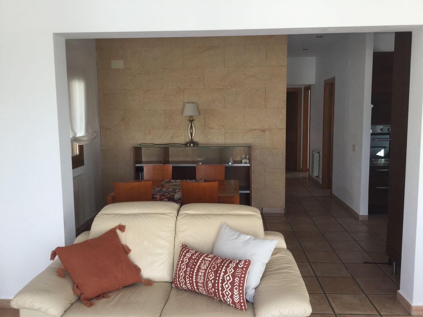 Salón con sofá de cuero crema y pared de piedra natural. El espacio abierto incluye un comedor y un pasillo con suelos cerámicos que conecta con la cocina.