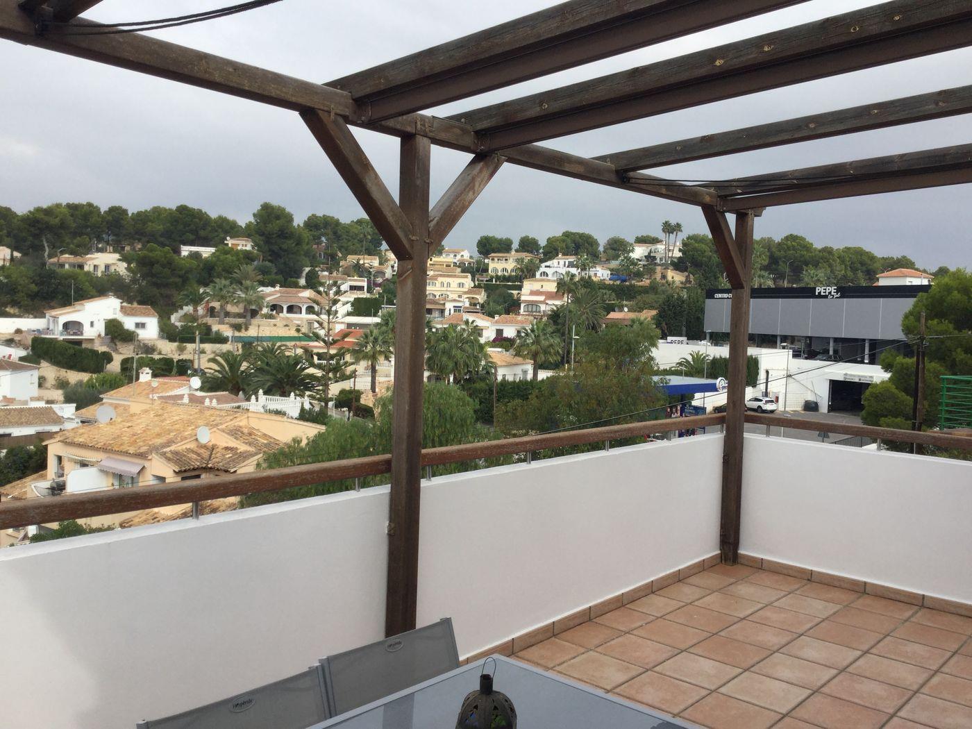 Terraza elevada con pérgola de madera y suelo de terracota, con vistas despejadas a una zona residencial mediterránea y vegetación local.