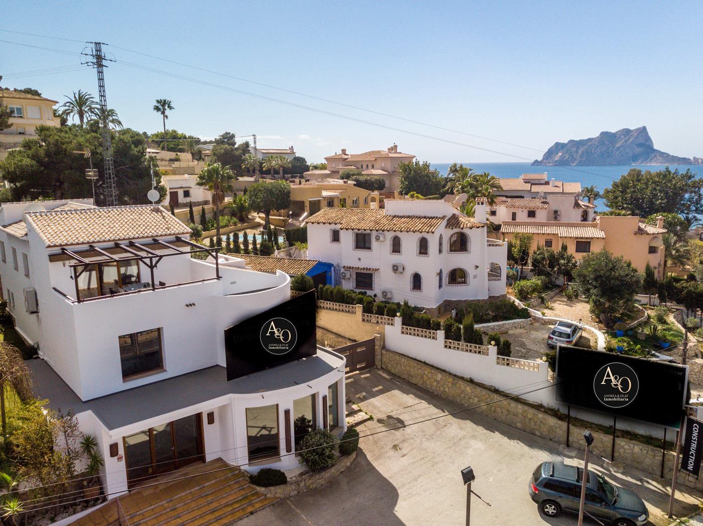 Vista elevada de una villa mediterránea blanca con fachada curva, terraza superior con pérgola de madera y vistas al mar con el Peñón de Ifach al fondo.