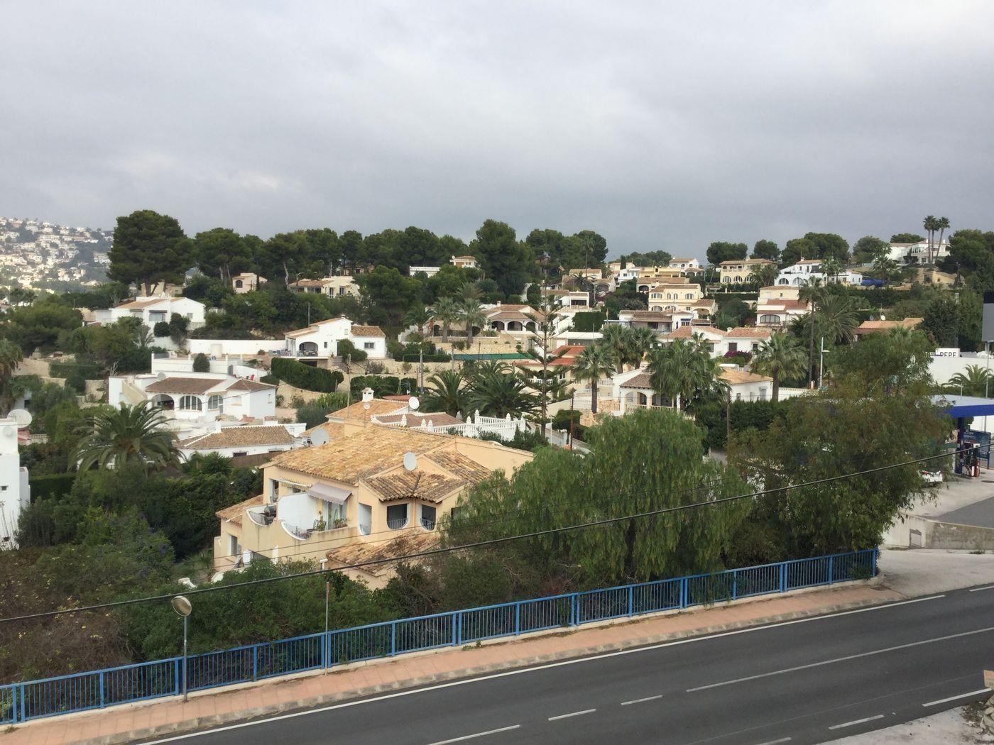Vista elevada de una zona residencial mediterránea con chalets blancos y tejados de terracota, rodeados de pinos y palmeras bajo un cielo nublado.