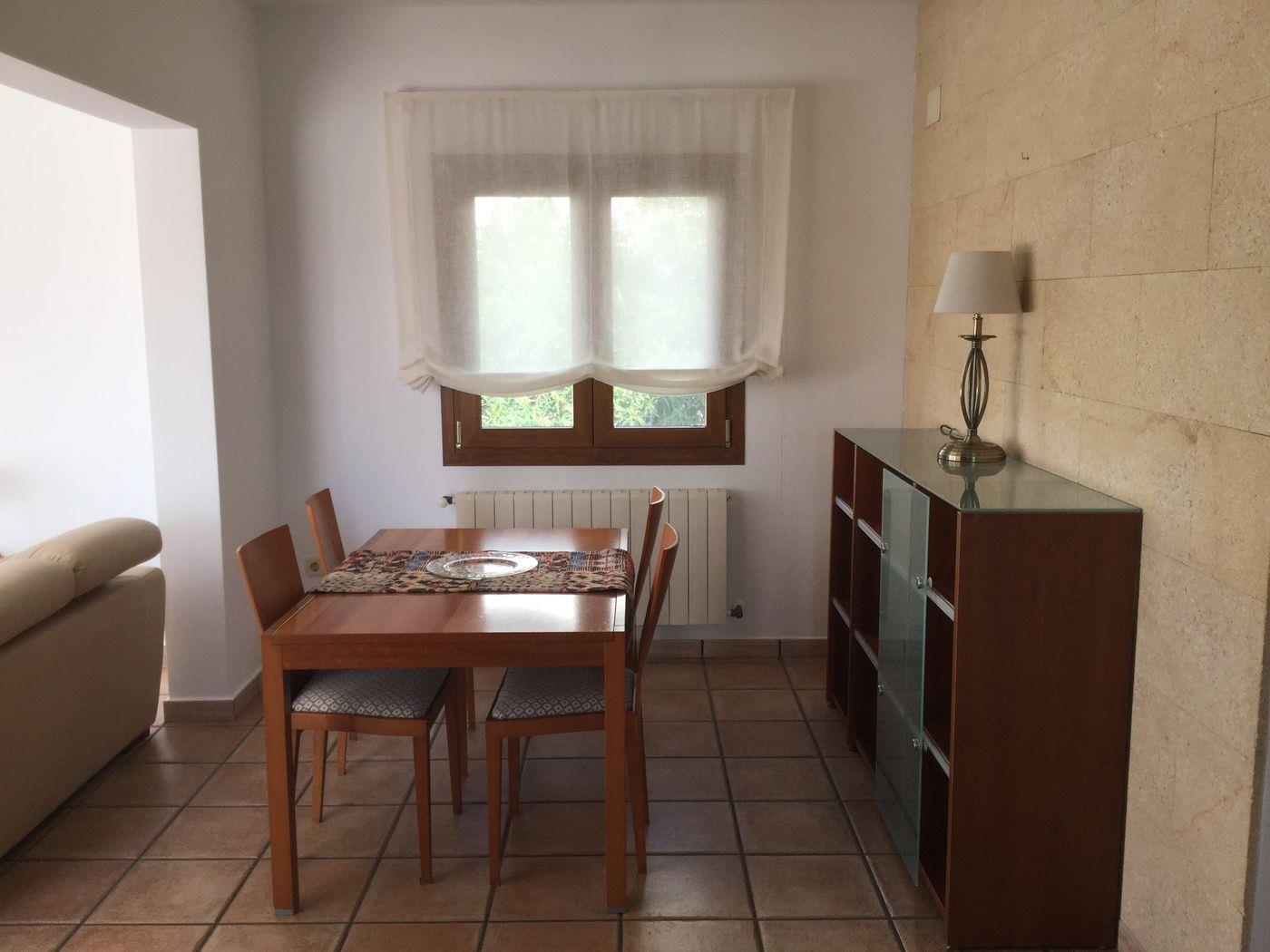 Zona de comedor con mesa de madera, suelos de baldosa cerámica y una pared de piedra natural. Incluye un aparador con vitrina y luz natural desde la ventana.
