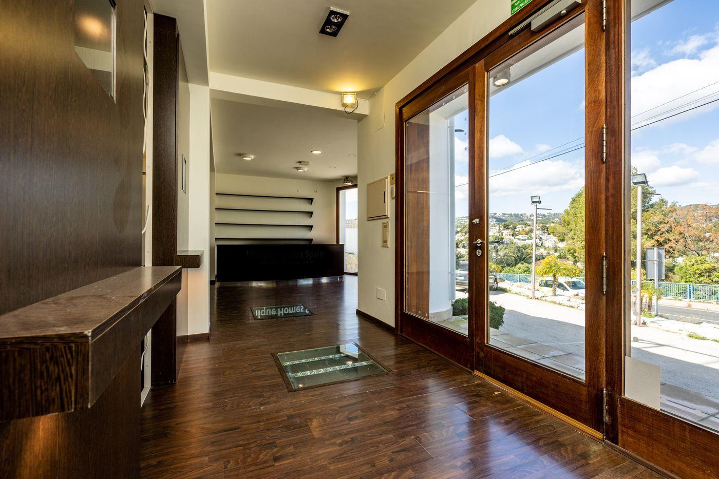 Zona de entrada con suelos laminados de madera oscura, panel de cristal integrado en el suelo y amplios ventanales de madera que muestran el mostrador de recepción.