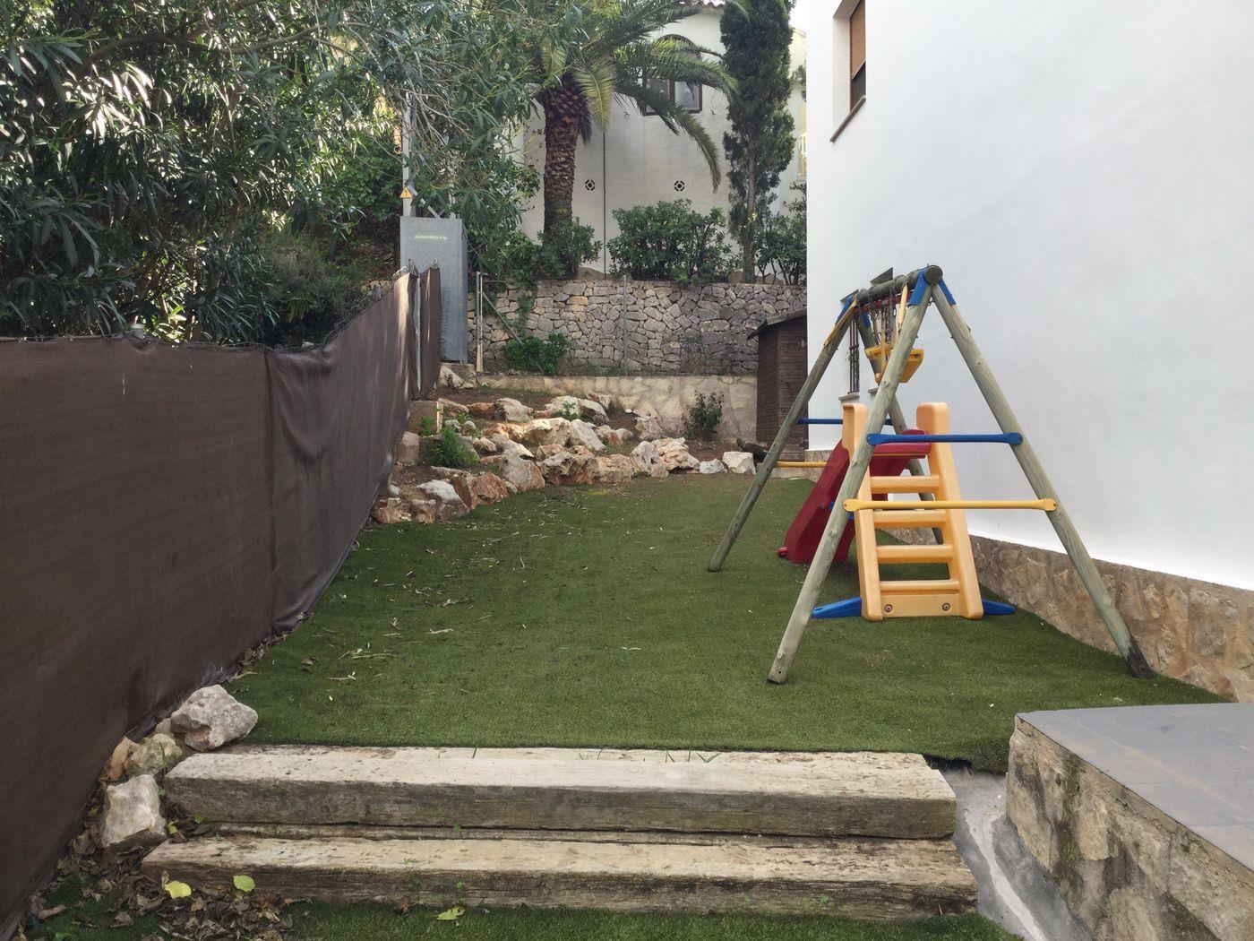 Zona infantil exterior con césped artificial, columpio de madera y peldaños de traviesas rústicas, delimitada por muros de piedra y vegetación mediterránea.