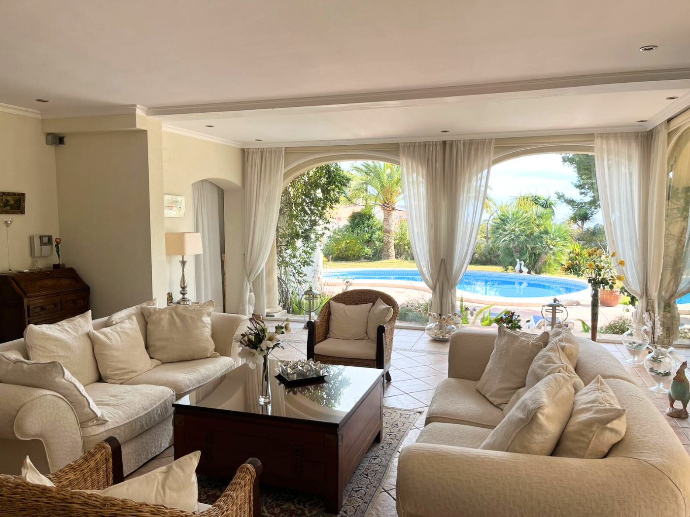 Amplio salón con sofás en tonos crema y sillones de mimbre, destacando sus grandes ventanales en arco con vistas directas a la piscina y al jardín mediterráneo.