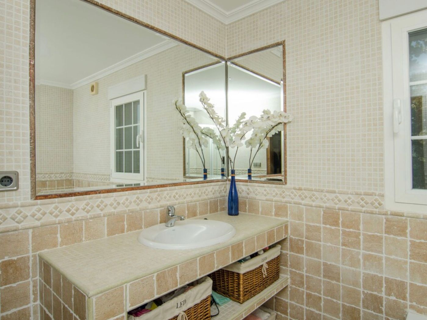 Baño con encimera de azulejos beige, lavabo encastrado y estantes con cestas de mimbre. Grandes espejos de pared a pared amplían visualmente la estancia.