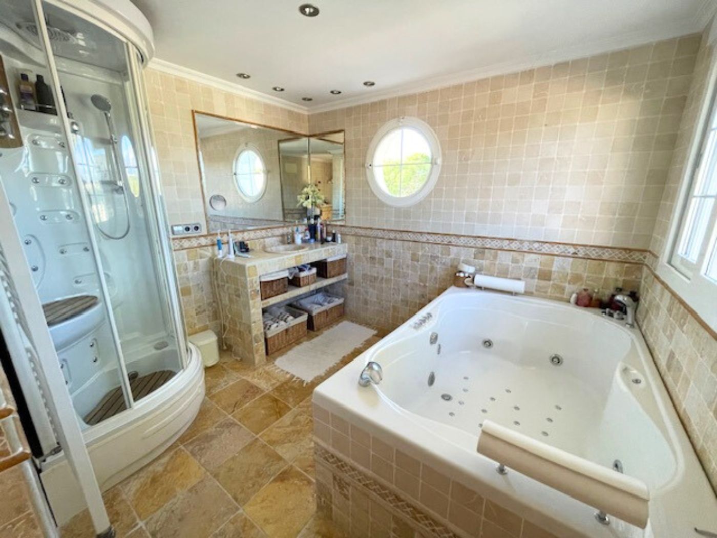 Baño equipado con bañera de hidromasaje, cabina de ducha acristalada y paredes revestidas en piedra con una ventana circular tipo ojo de buey.