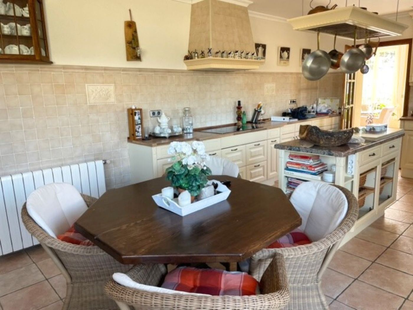 Cocina con mesa octogonal de madera, sillas de mimbre y una isla central con encimera de mármol, complementada por armarios clásicos y suelos cerámicos.