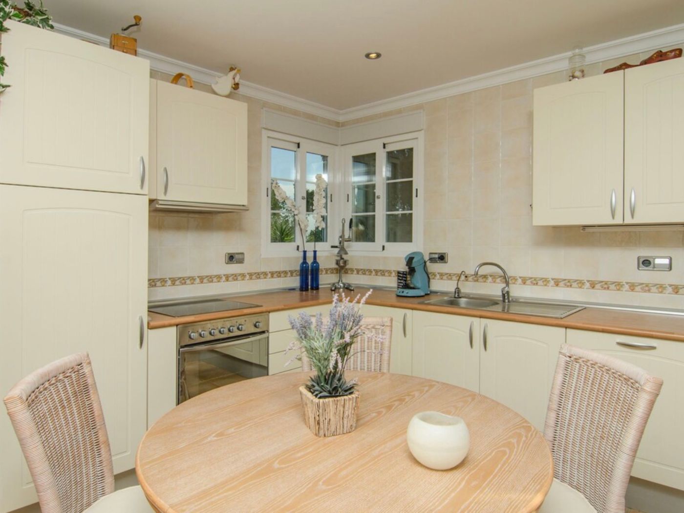 Cocina con mobiliario crema, encimeras de madera y zona de comedor con mesa redonda y sillas de mimbre, complementada por paredes alicatadas.