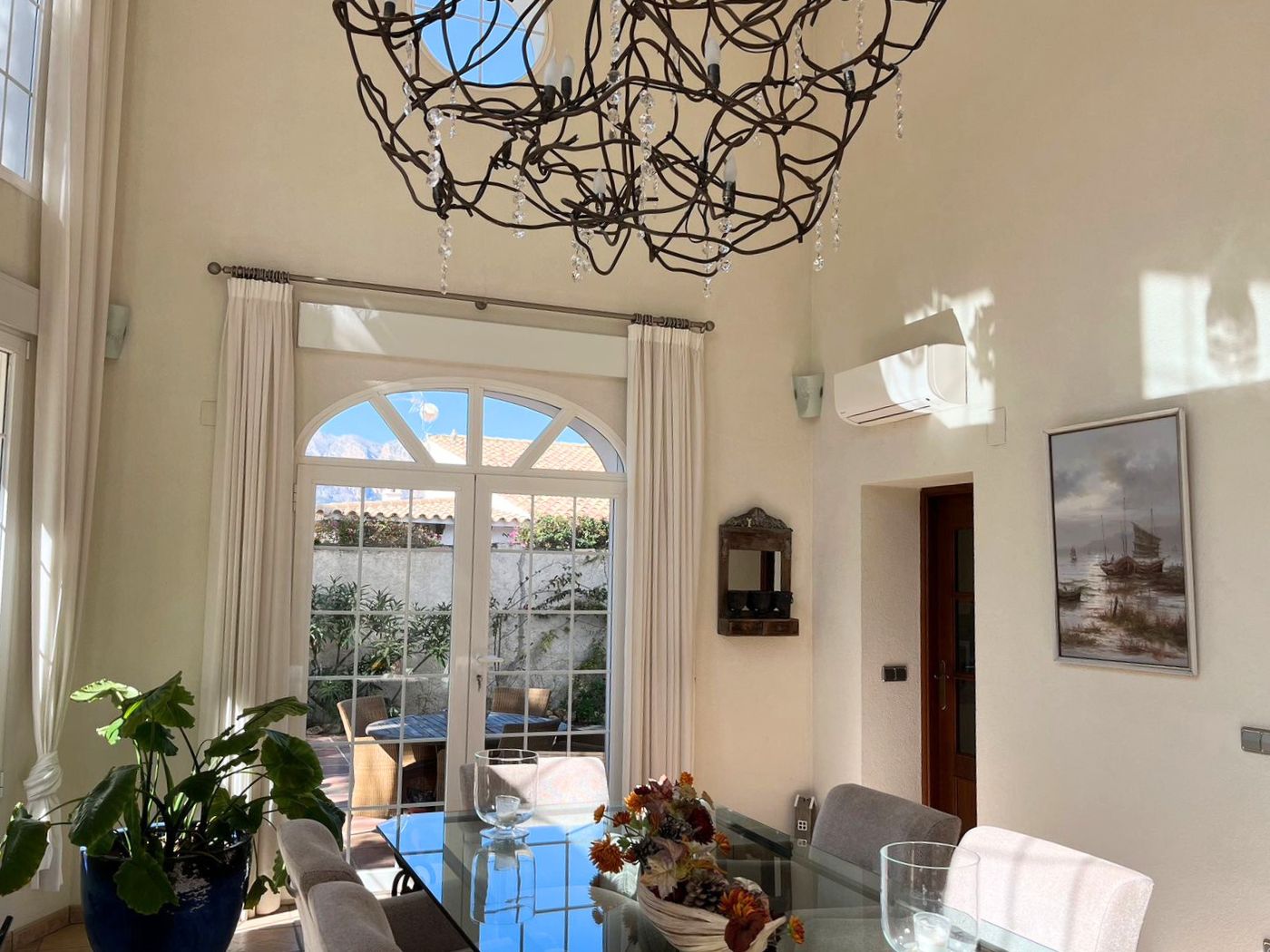 Comedor de techos altos con mesa de cristal y lámpara de hierro forjado. Puertas de arco conectan el interior con una terraza privada ajardinada.