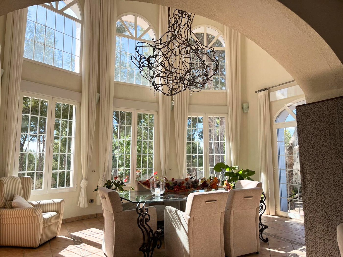 Comedor luminoso con techos de doble altura, grandes ventanales de arco, mesa de cristal con base de hierro forjado y una lámpara escultural de diseño.