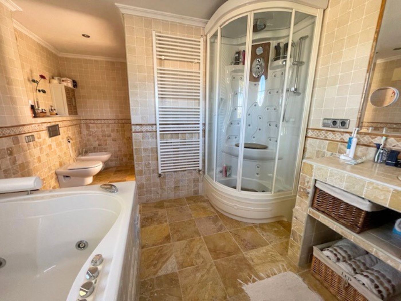 Cuarto de baño con cabina de ducha de hidromasaje, bañera amplia y revestimientos de piedra natural. Dispone de radiador toallero y mobiliario con cestas de mimbre.