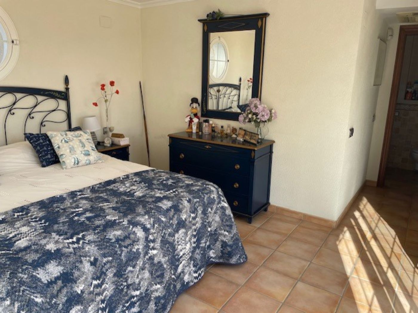 Dormitorio con cabecero de forja, cómoda azul marino con espejo a juego y suelos de baldosa de terracota. La estancia incluye una ventana circular y luz natural.