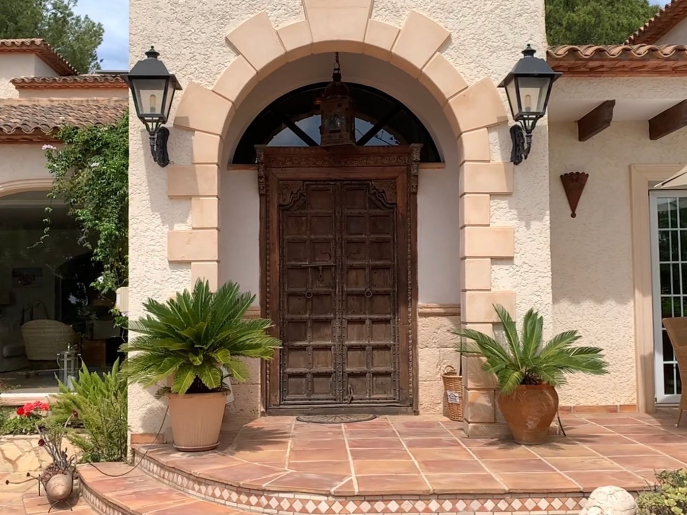 Entrada con puerta doble de madera tallada antigua bajo un arco de piedra, flanqueada por faroles clásicos y palmeras cicas en macetas sobre suelo de terracota.