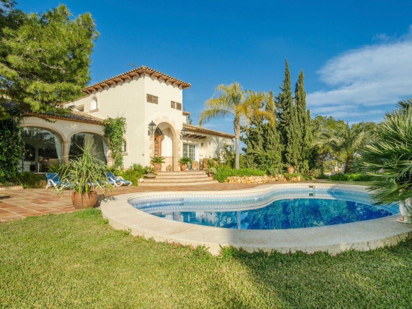 Exterior de villa mediterránea con piscina de formas curvas, terraza de terracota y fachada de estuco blanco con arcos tradicionales y jardín consolidado.